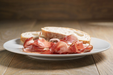 speck slices on white plate with ciabatta on old wood table