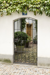 Blick durch ein Metallgitter in einen hübschen kleinen Innenhof eines Hauses mit weissen Wänden, Kopfsteinpflaster, Pflanzen und einer braunen Holztür.