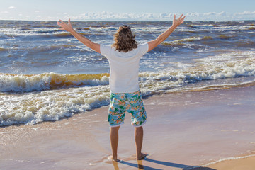 Young man at the seaside with waves. He breathes the fresh air and feels the freedom in sunny summer day.