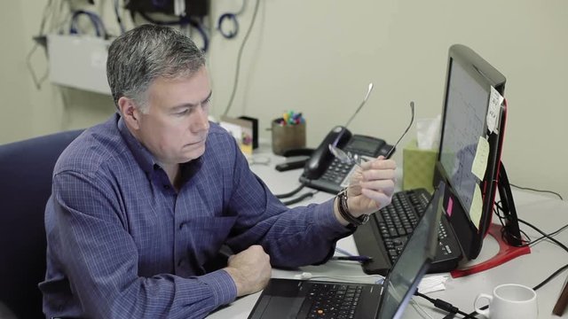 A mature network administrator or technician in office reviews data on two computer screens then smiles