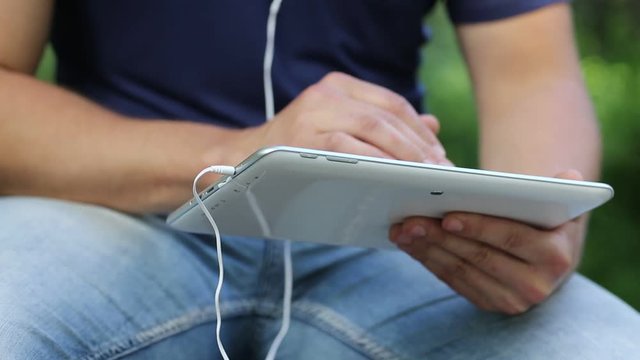 Man looks the photos in his tablet computer. Man holds in hands white tablet pc. Closeup of man hands with white tablet pc and earphones. Man looks and flips through the photos in tablet computer
