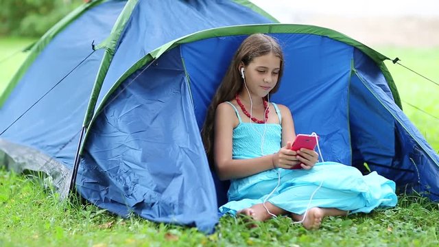 Beautiful girl sits on the grass near blue tourist tent and communicates via smartphone. Beautiful girl in light blue dress with red smartphone sits on the grass. Pretty girl talks on phone via skype