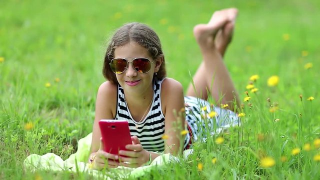 Attractive girl in sunglasses with red smartphone lies on the grass. Pretty girl playing the game on his smartphone. Little girl in glasses lies on the green grass and uses cell phone