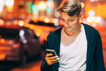 Handsome young man using smartphone while walking in night city streets. Smiling guy reading message outdoor