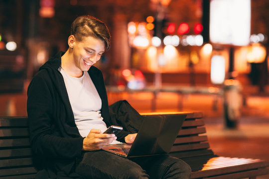 Smiling guy reading message on smartphone, while he working on laptop outside. Young male freelancer working in city street in night time, blurred lights background, free space