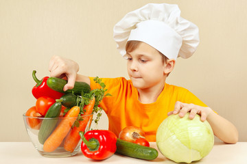 Little boy in chefs hat chooses vegetables for salad at the table