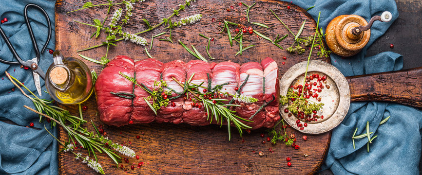 Raw roast beef  with herbs tied with a rope with cooking ingredients, oil  and spices on rustic background, top view, banner