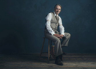 Confident retro 1920s fashion man sitting on wooden chair in emp