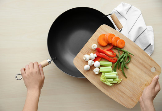 Female hands holding cutting board with vegetables and pan on wooden table, top view