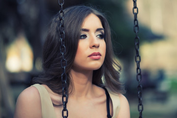 Outdoor portrait of beautiful young woman. She siting on park swing and looking at side.