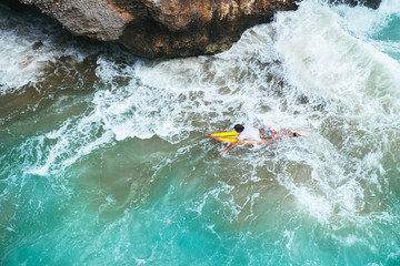 Outdoors aerial view of surfer swimming on a board near huge blue ocean wave and dangerous cliff in Bali, Indonesia