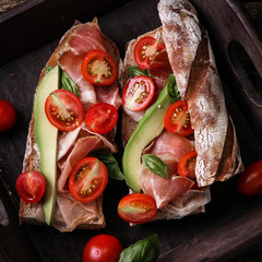 Ciabatta sandwich with prosciutto,avocado,cherry tomatoes and basil on dark background