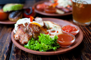 Barbecued pork or lamb meat shish kebab grill with salad, vegetables, glass of beer and sauce on clay dish. Vintage wooden table background.