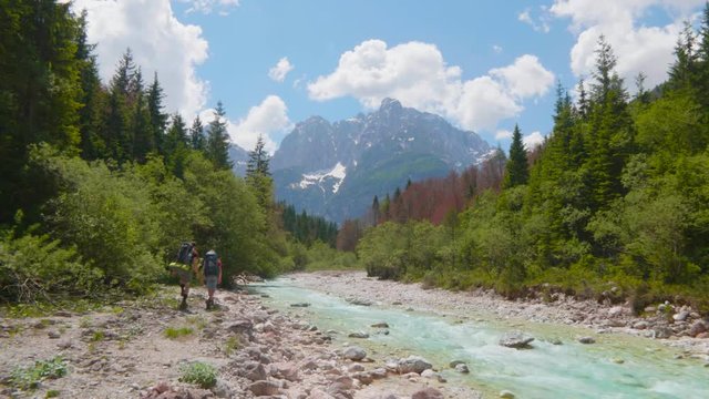 Mid air tracking shot of two hikers walking along a beautiful emerald river with high mountains and the blue sky in the background.
