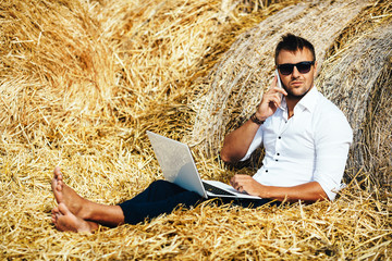 Business man looks beautiful works with a laptop and talking on the phone sitting in haystack.