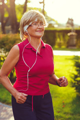 Portrait of elderly woman running with headphones and smartwatches in the park in evening sunset