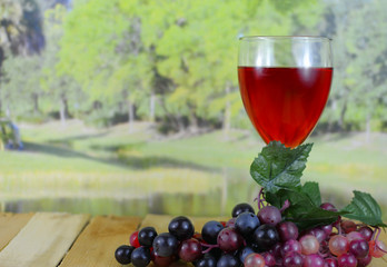 A glass of dark pink wine sitting on a wooden table with grapes with a blurred background of a tree lined lake in the sunshine. There is condensation on the glass and reflections from the lights.