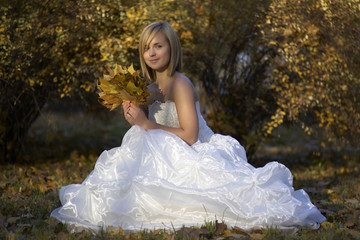 Fototapeta premium Happy Beautiful young bride in white dress sitting in autumn park among fallen leaves/Gorgeous blonde bride in wedding dress with fallen leaves bouquet posing in sunny autumn park