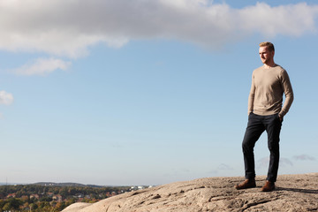 A handsome blonde man wearing a beige pullover shirt, standing high up on a mountain overlooking...
