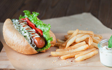 Big tasty hot dog with sausage and vegetables with sauce and French fries on the parchment on the wooden background. hot dogs to gourmet.