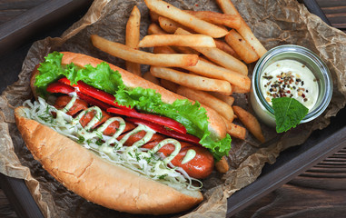 Big tasty hot dog with sausage and vegetables with sauce and French fries on the parchment on the wooden background. hot dogs to gourmet.