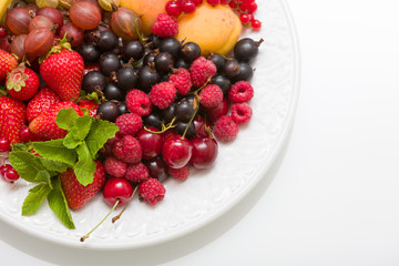 Assortment of juicy fruits on white plate and white table background. Strawberries, blueberries, raspberries, gooseberry, blackcurrant decorated with mint for summer dessert or snack.
