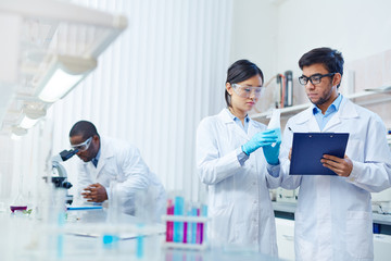 Concentrated Asian female laboratory scientist in safety goggles holding flask with blue liquid showing it to Latin-American colleague writing down results. African-American scientist in background.