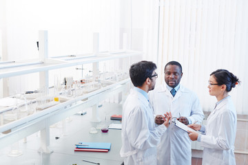 High angle shot of pleased male African-American, male Latin-American and female Asian laboratory scientists in lab coats and glasses discussing scientific research. - obrazy, fototapety, plakaty