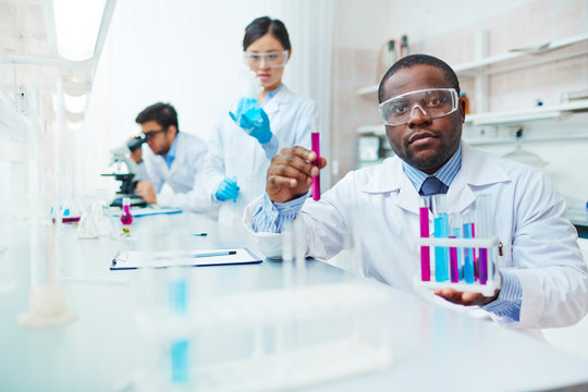 Waist up of male African-American scientist in lab coat and safety goggles looking at camera holding test tubes, female Asian colleague examining flask, male Latin-American scientist in background.