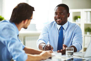 Smiling African-American businessman in shirt and necktie sitting in office talking about work with Caucasian businessman