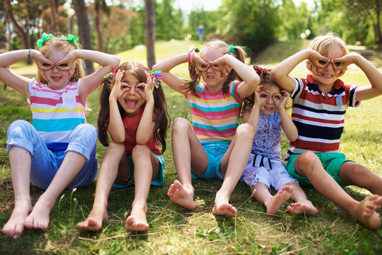 Portrait of four smiling little girls with ponytails and one little blond boy sitting on grass in green park on sunny day holding fingers near eyes like glasses, pulling faces to camera.