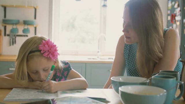 Mother is smiling while talking to her little daughter who is colouring a picture on the kitchen table. Slow mo