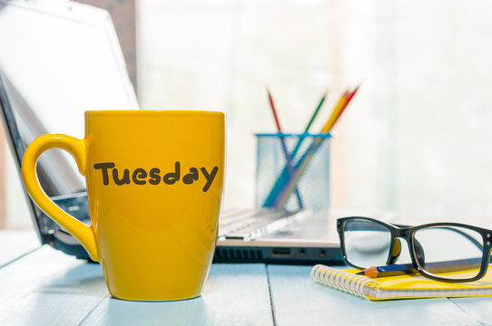 Tuesday written on yellow coffee or tea cup at wooden boards table, workplace, office sunlight morning background