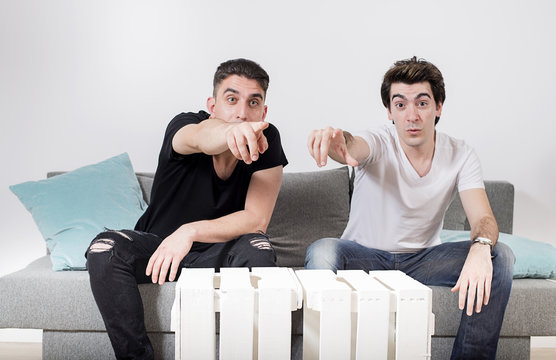Two male friends sitting on a gray sofa with cushions pointing to the camera