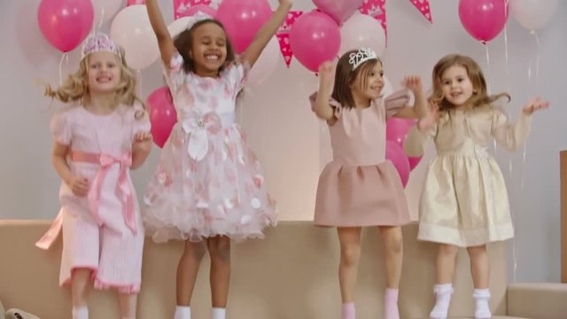Four carefree little girls dressed like little princesses jumping on sofa together in the living room at birthday party, smiling and waving
