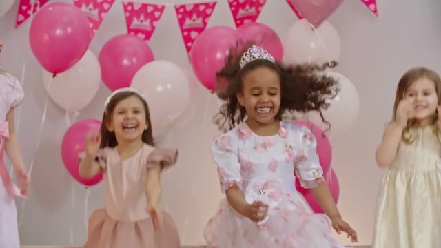 Slow motion shot of four little girls in dresses and tiaras jumping together on sofa decorated with balloons 