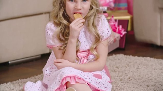 Tilt up of blonde little girl with beautiful long hair dressed in fancy dress and crown sitting on the carpet and eating cookie