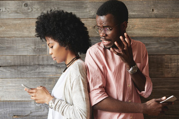 Portait of annoyed African man in glasses angry with his attractive girlfriend who is absorbed in...