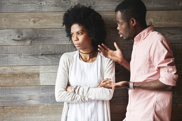 Young displeased black male gesturing in indignation, arguing with his stylish girlfriend, who is...