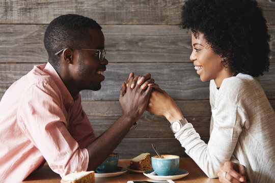 Dark-skinned young couple sitting at restaurant: black man in glasses holding his girlfriend's hands, declaring his love to her or proposing on their anniversary day, both looking happy and cheerful