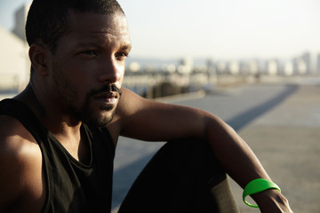 Cropped shot of exhausted African male athlete having rest outside after intensive workout sitting at sidewalks at sea thinking about his future career, looking serious, concentrated and thoughtful