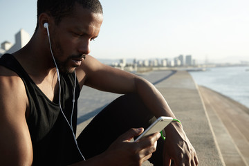 Handsome dark-skinned jogger wearing black sleeveless top listening to music with headphones,...