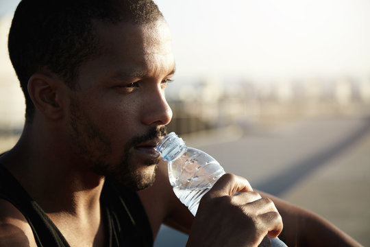 Healthy lifestyle concept. Profile portrait of black man with athletic body sitting on pavement in morning sun after training exercises in open air, holding bottle, drinking water, looking far away