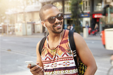 People, technology and communication. Charismatic young African traveler having walk on city street, talking to his friends on mobile phone using handsfree, laughing at jokes, looking happy and glad