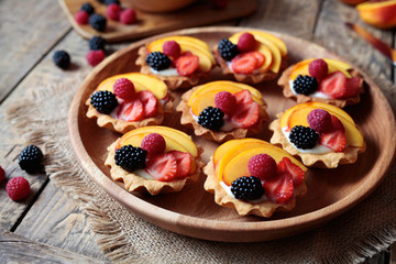 Fruit dessert tartlets with vanilla custard and fresh raspberries, blackberry and peach served on wooden tray with baking forms, kitchenware on the old wooden table. Dark rustic style.