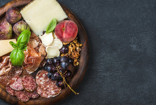 Italian antipasti snack for wine. Prosciutto di Parma, salami, cheese variety, figs, grapes, peach, walnuts and fresh basil on wooden serving tray over dark grunge background. Top view, copy space