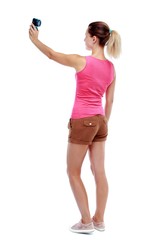 back view of standing young beautiful woman and using a mobile phone. Isolated over white background. Sport blond in brown shorts makes mobile camera selfie.