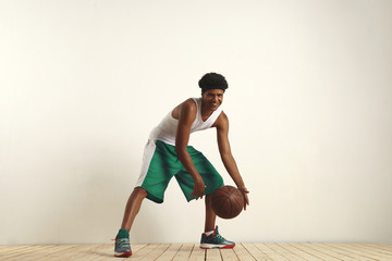 Smiling young basketball player in white and green sportswear dribbling looking into camera against white background