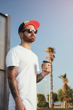 Young man with beard and tattoos in unlabeled white t-shirt with a coffee cup against blue sky and palm trees