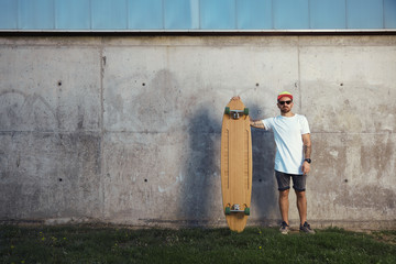 Serious looking surfer with beard, tattoos and sunglasses standing next to his longboard looking directly into camera